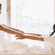 Kobieta w białym stroju delikatnie rozciąga ramię leżącej osoby na białym materacu przy jasnym oknie, tworząc relaksującą atmosferę.