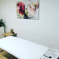 Biały stół do masażu w jasnym pomieszczeniu, nad stołem obraz z motywem kwiatowym, obok stolik z kosmetykami i roślina doniczkowa.