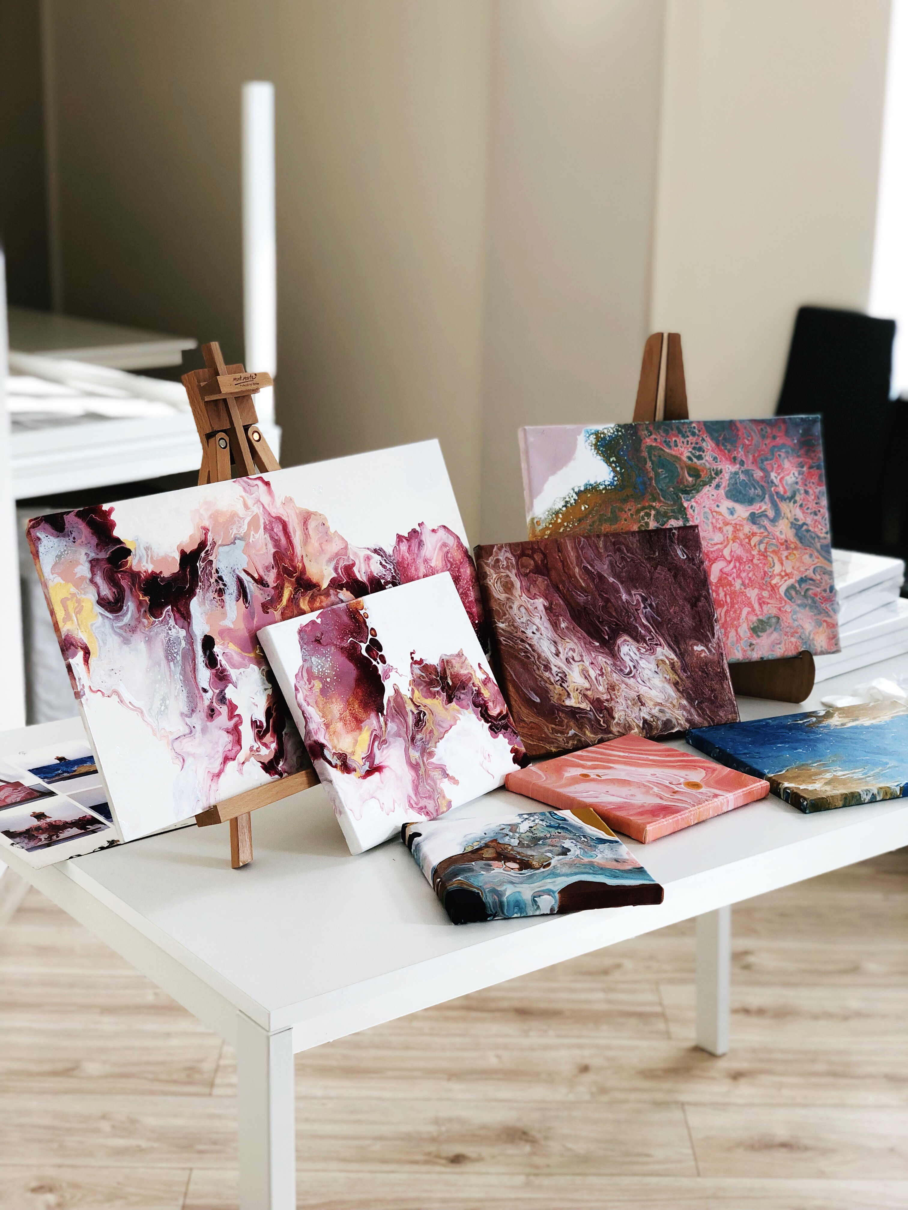 Seria obrazów akrylowych w technice fluid art na białym stole, niektóre na małych drewnianych sztalugach, prezentujących abstrakcyjne wzory w odcieniach różu, brązu, błękitu i złota.