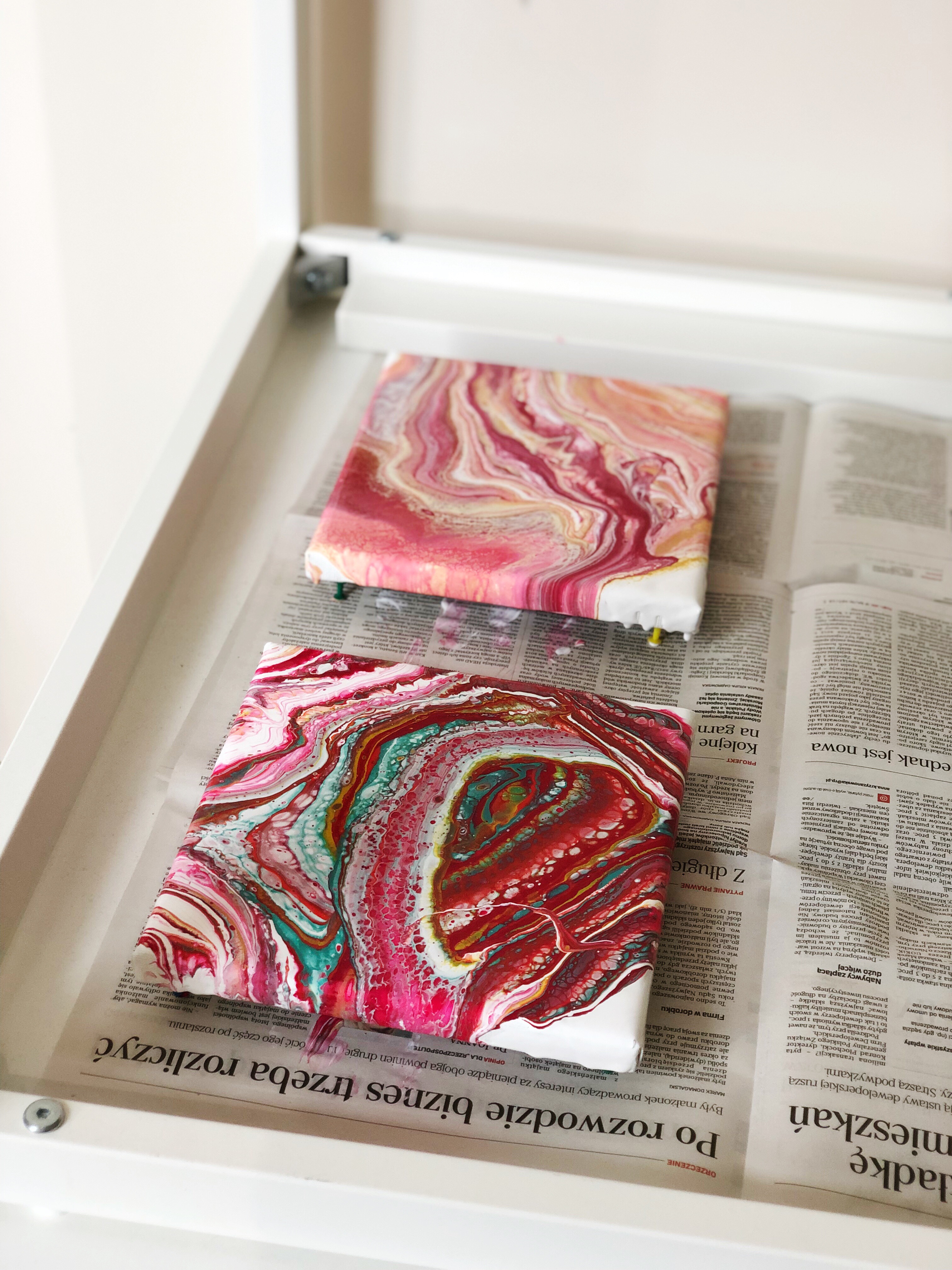 Dwa kwadratowe obrazy akrylowe w technice pouring art, jeden w odcieniach różu i bieli, drugi z dodatkiem zieleni i brązu, leżące na gazecie w białej szufladzie.