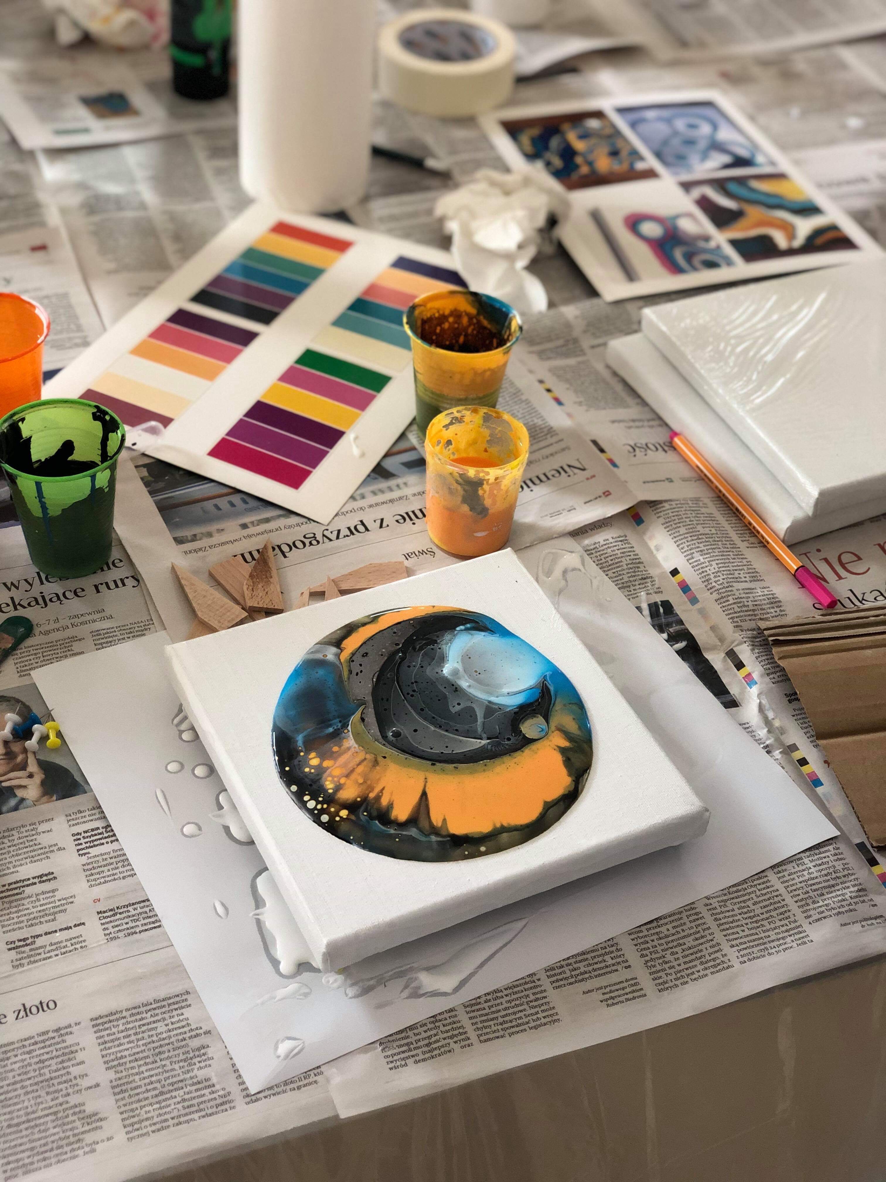 Warszawa: Praca w toku nad abstrakcyjnym obrazem na płótnie techniką pouring art, widoczne kubeczki z farbami, paleta kolorów i gazety zabezpieczające powierzchnię.