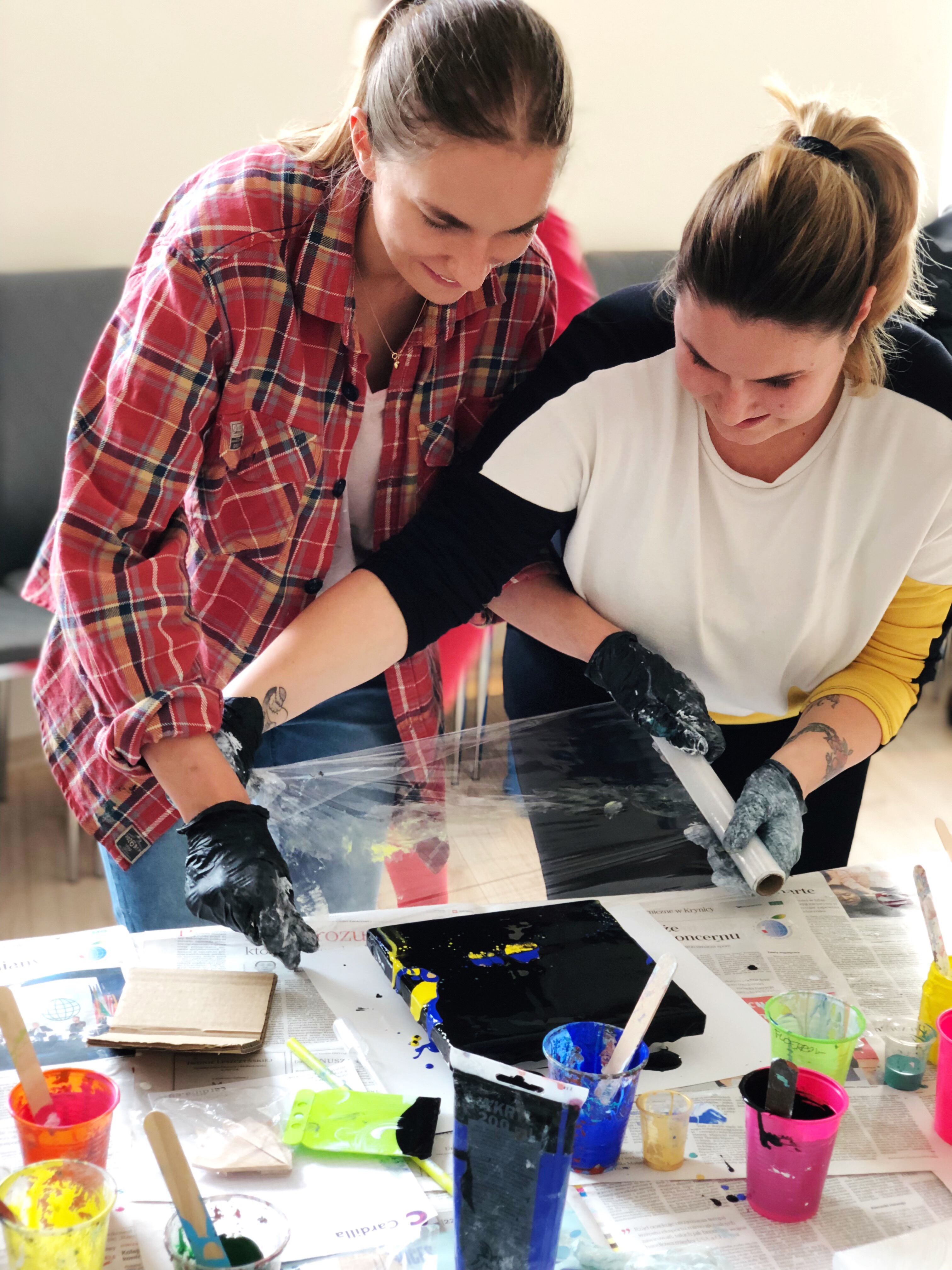 Dwie kobiety w rękawicach ochronnych pracują nad obrazem z wykorzystaniem techniki pouring art, widoczne farby w kubeczkach i narzędzia.