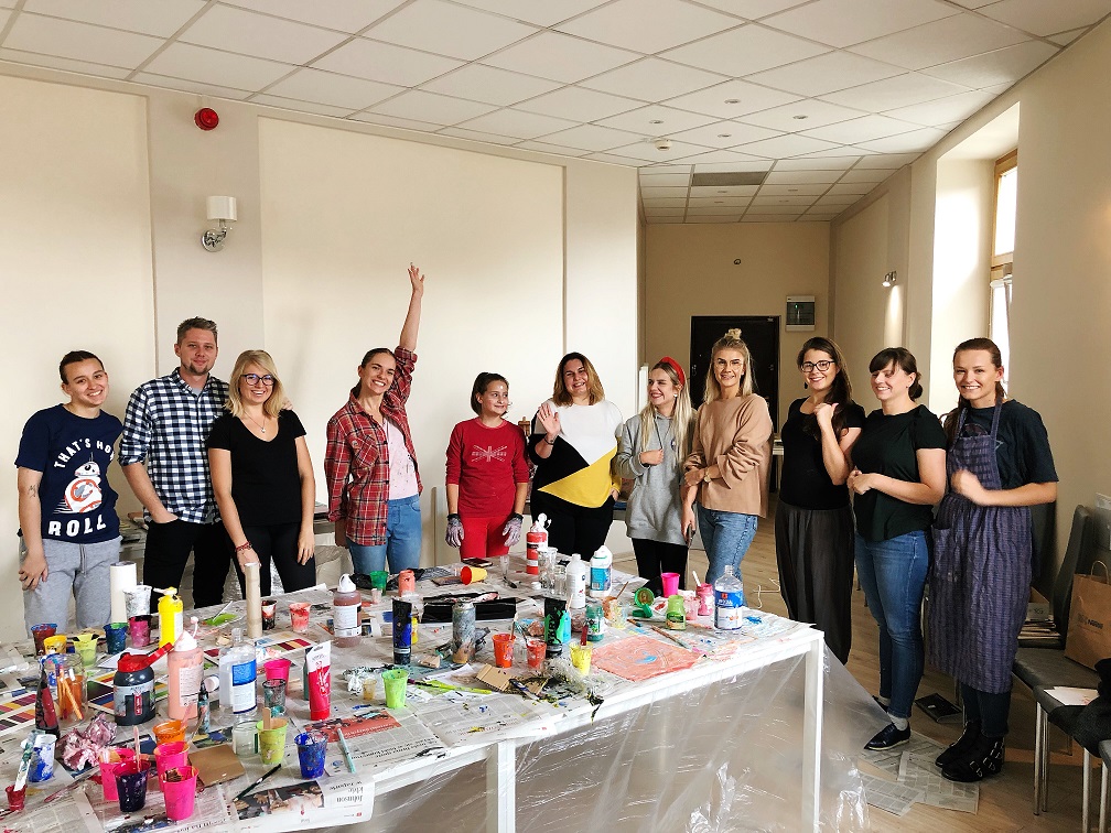 Grupa osób uczestniczy w warsztatach artystycznych, stół pokryty farbami i pędzlami, jedna osoba z podniesioną ręką, jasne wnętrze.