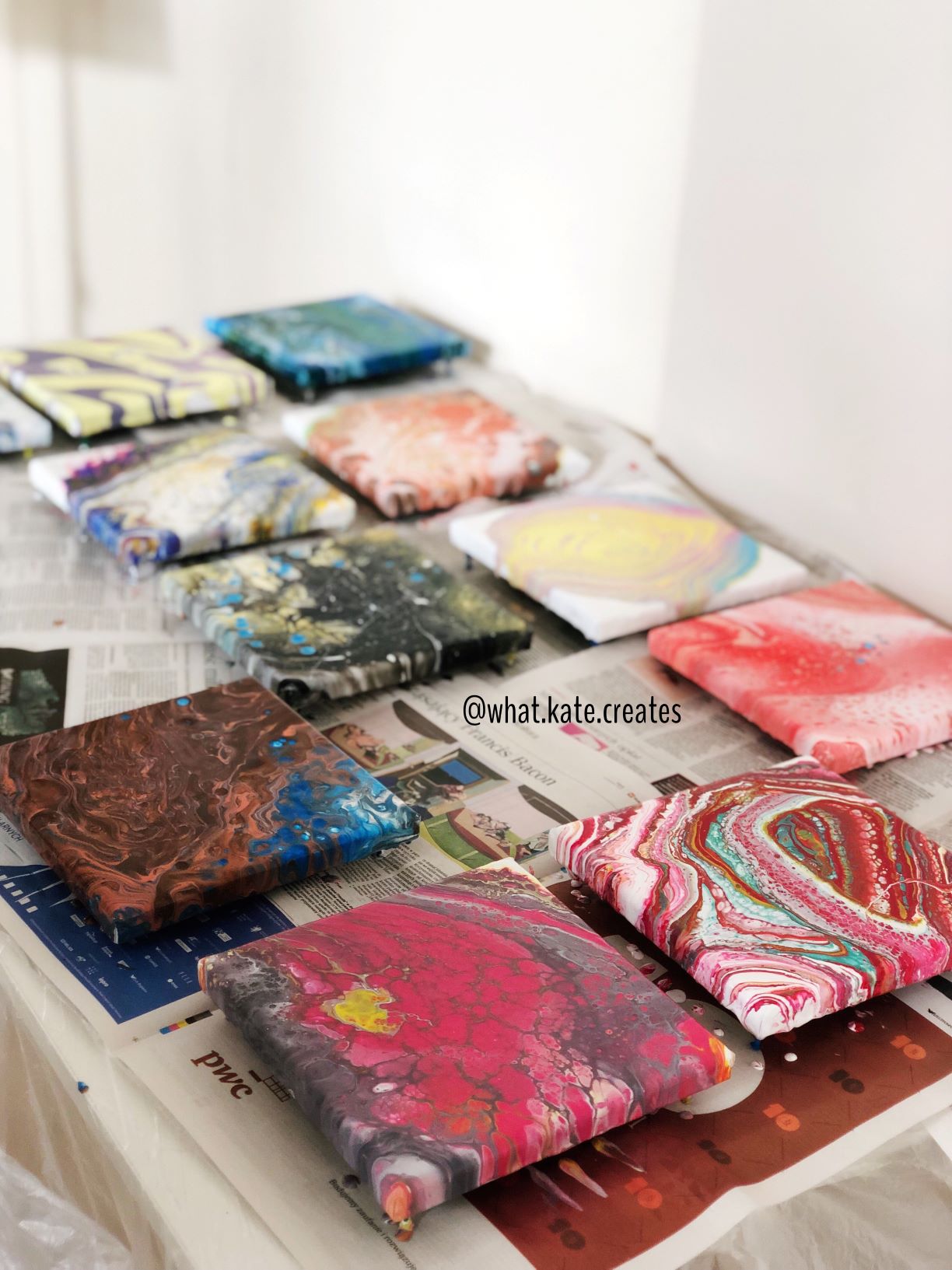 Seria kwadratowych płócien z abstrakcyjnymi wzorami wykonanymi techniką pouring art, ułożonych na gazecie z widocznym logo PwC.