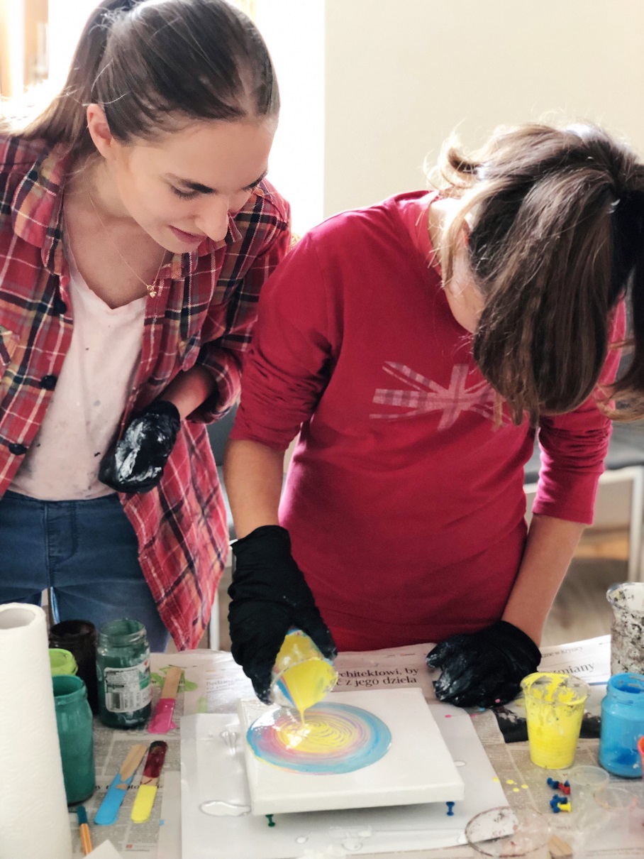 Dwie kobiety tworzą abstrakcyjny obraz techniką wylewania farb na płótno, używając czarnych rękawic ochronnych, z widocznymi pojemnikami z farbami i gazetą jako podkładem.