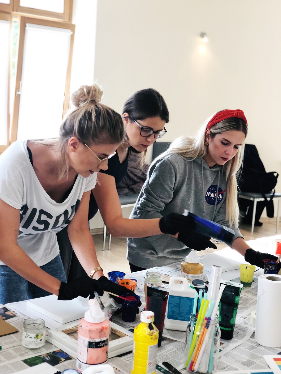 Trzy kobiety w czarnych rękawicach malują farbami akrylowymi na płótnach podczas warsztatów artystycznych. Używają różnych kolorów i technik w jasnym, słonecznym pomieszczeniu.