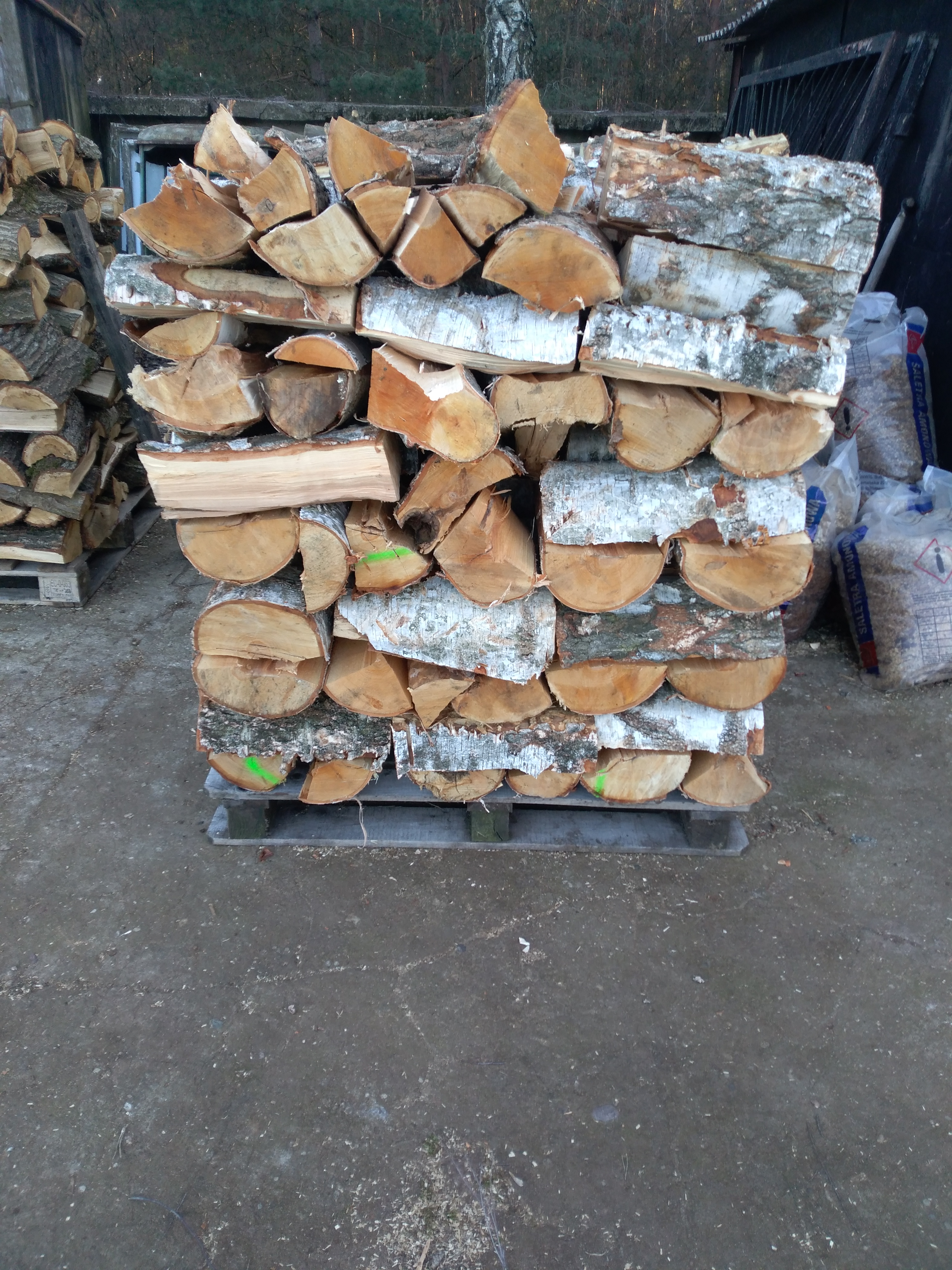 Ułożone na palecie pocięte kawałki drewna kominkowego, głównie brzozowego, z widocznymi zielonymi oznaczeniami na niektórych kawałkach, w tle worki z pelletem i fragment budynku.