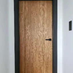 Zamknięte drzwi wewnętrzne z jasnego drewna o wyraźnym usłojeniu, z czarną ościeżnicą i minimalistyczną klamką, zamontowane w białej ścianie, z widocznym panelem sterowania oświetleniem po prawej...