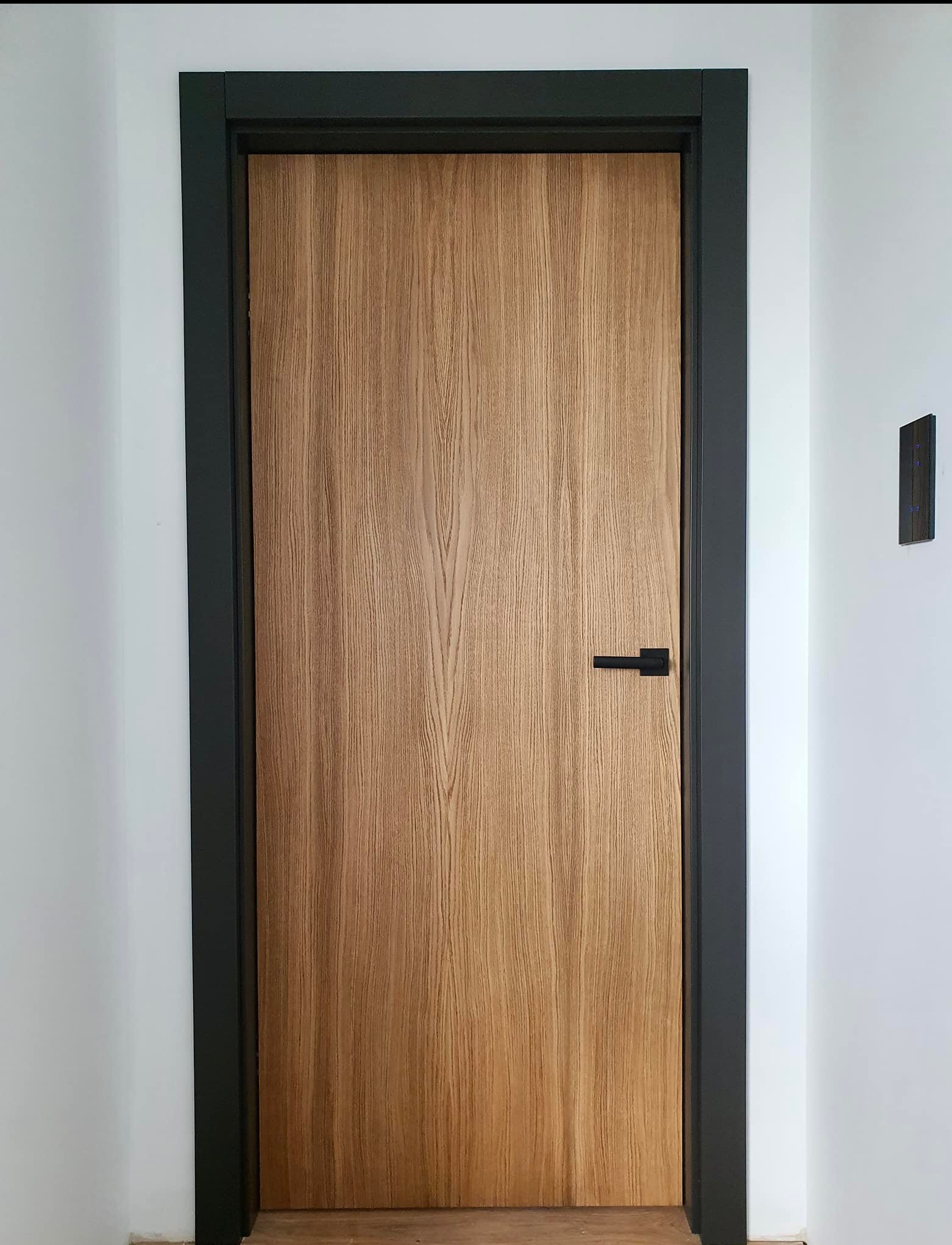 Zamknięte drzwi wewnętrzne z jasnego drewna o wyraźnym usłojeniu, z czarną ościeżnicą i minimalistyczną klamką, zamontowane w białej ścianie, z widocznym panelem sterowania oświetleniem po prawej...