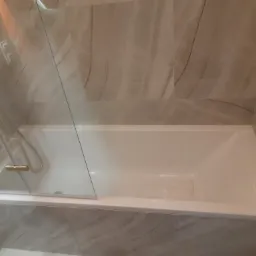 Biała wanna z zamontowaną szklaną ścianką działową z elementami w kolorze złotym, otoczona płytkami imitującymi marmur.