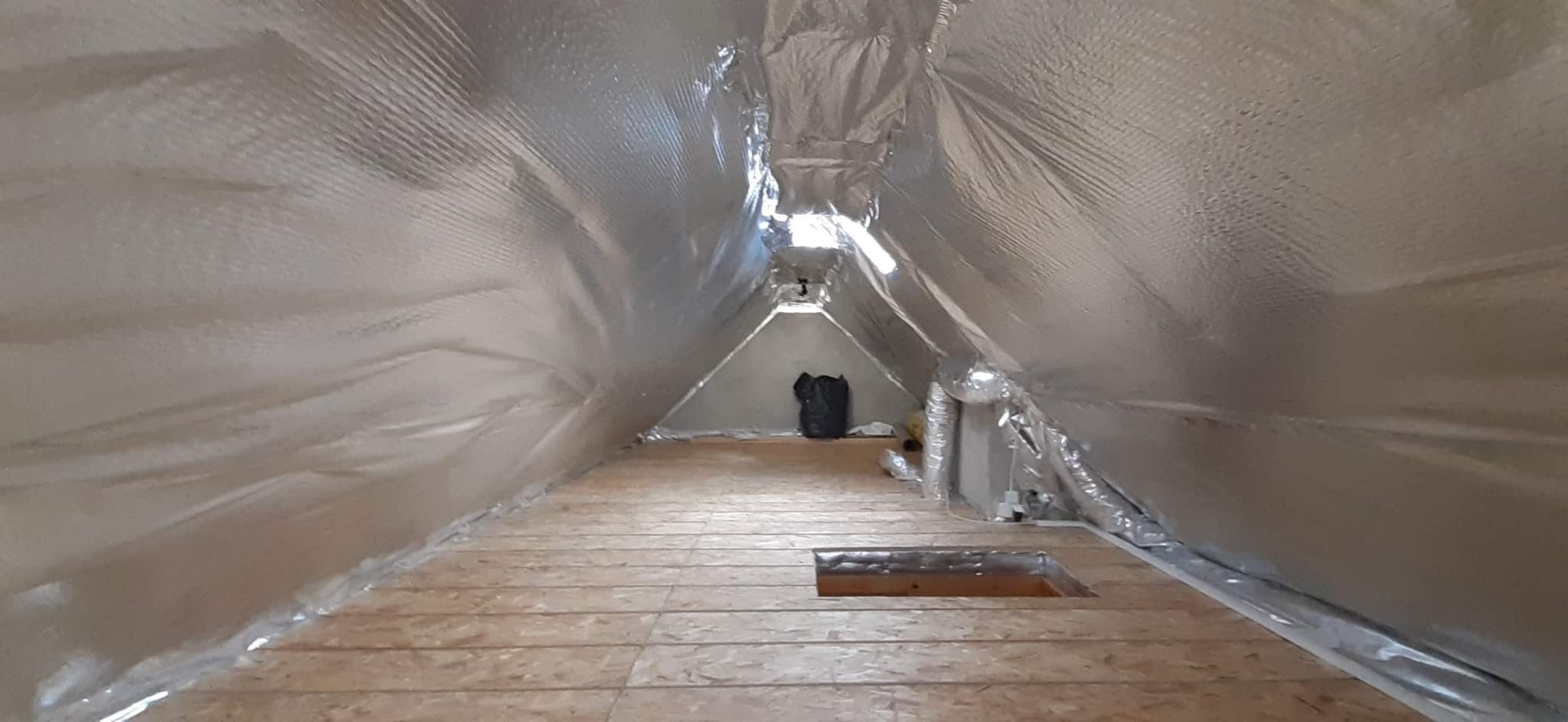 Izolacja poddasza folią aluminiową, widoczne deski podłogowe i otwór w podłodze.