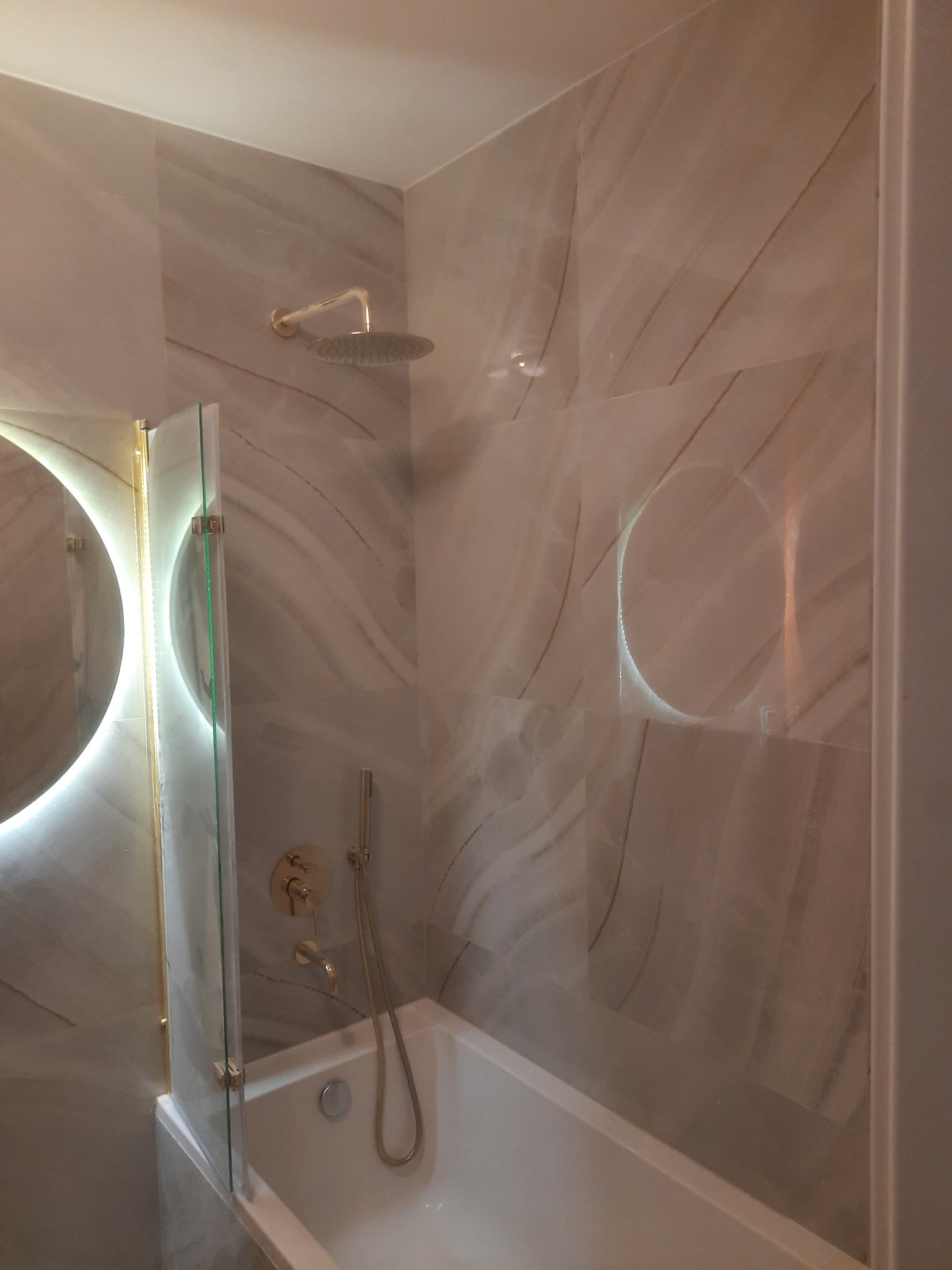 Nowoczesna łazienka z beżowymi płytkami imitującymi marmur, okrągłe lustro z podświetleniem LED, złota armatura, szklany parawan nawannowy i biała wanna.