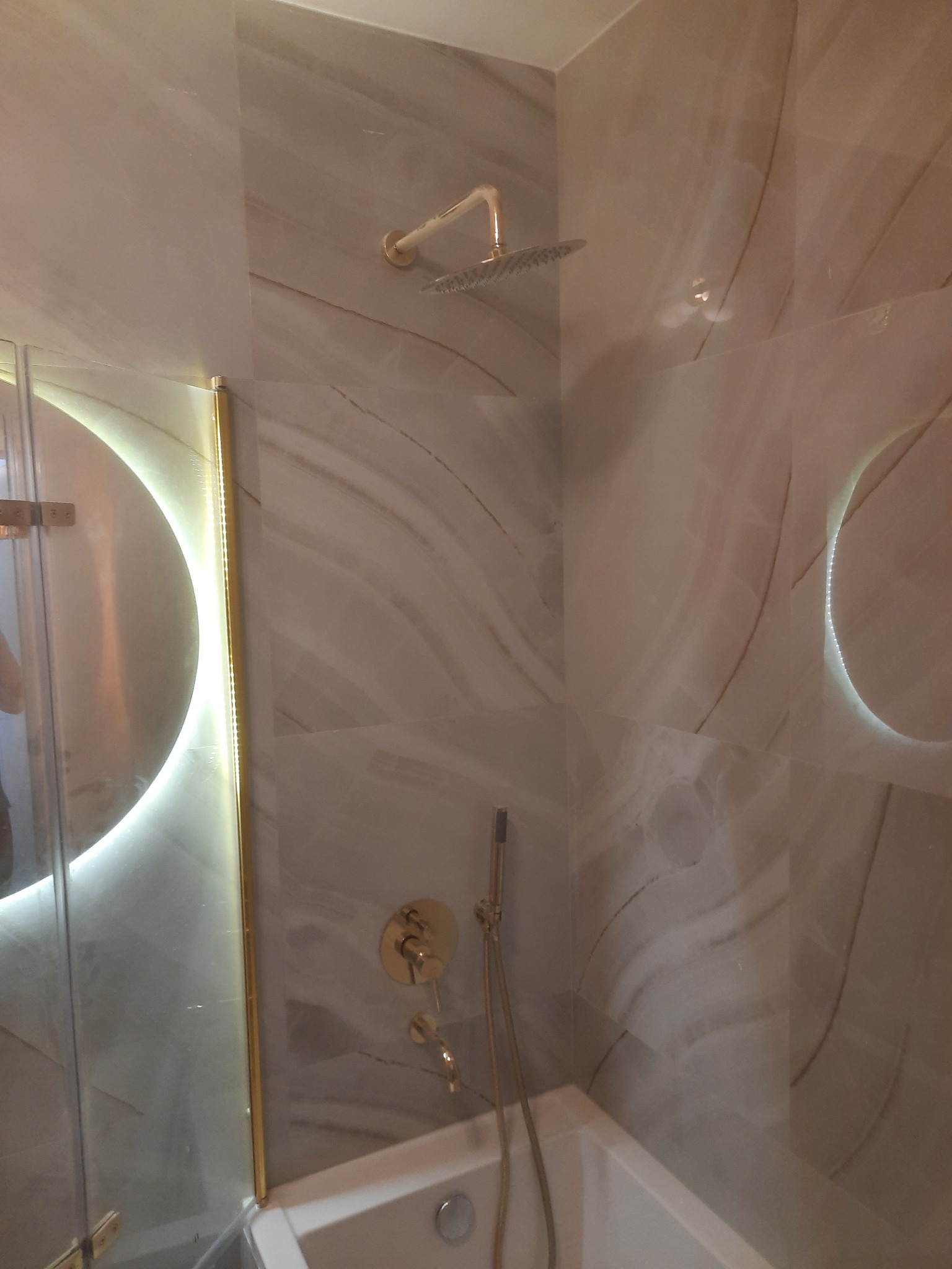 Wykończona łazienka z beżowymi płytkami imitującymi marmur, złotą armaturą, okrągłym lustrem z podświetleniem LED oraz białą wanną.