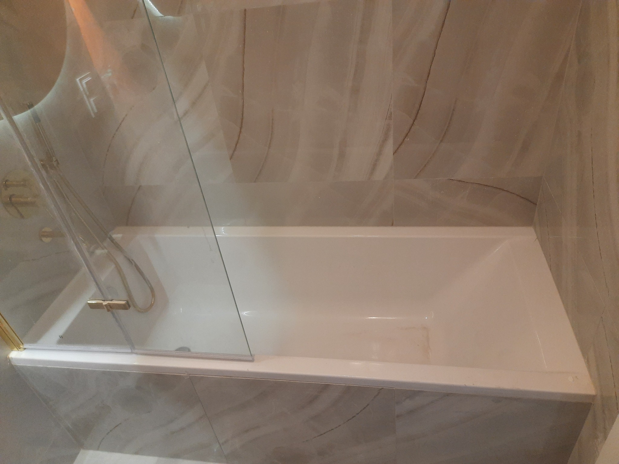 Biała wanna z zamontowaną szklaną ścianką działową z elementami w kolorze złotym, otoczona płytkami imitującymi marmur.