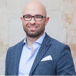 Portret uśmiechniętego, łysiejącego mężczyzny w okularach, w niebieskiej koszuli i szarej marynarce z poszetką, na tle ściany z jasnych kamiennych bloków.