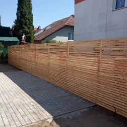 Nowoczesne ogrodzenie z poziomych desek z jasnego drewna, ułożonych z przerwami, na tle kostki brukowej i domu jednorodzinnego z ogrodem.