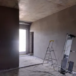 Wnętrze w trakcie prac wykończeniowych: ściany pokryte świeżo nałożoną warstwą zaprawy, widoczna drabina i sprzęt do natrysku, surowy sufit betonowy, przewody elektryczne na podłodze.