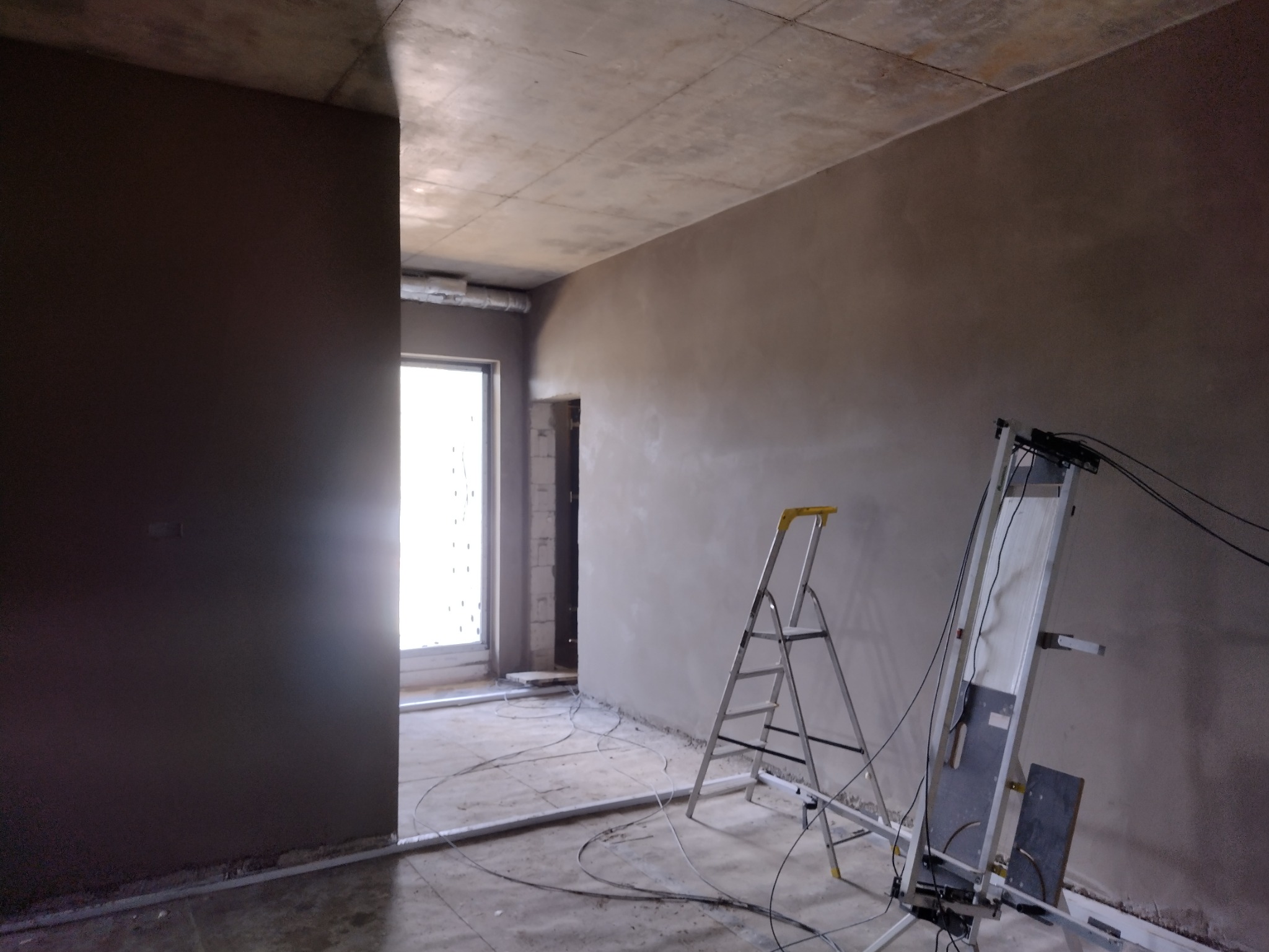 Wnętrze w trakcie prac wykończeniowych: ściany pokryte świeżo nałożoną warstwą zaprawy, widoczna drabina i sprzęt do natrysku, surowy sufit betonowy, przewody elektryczne na podłodze.