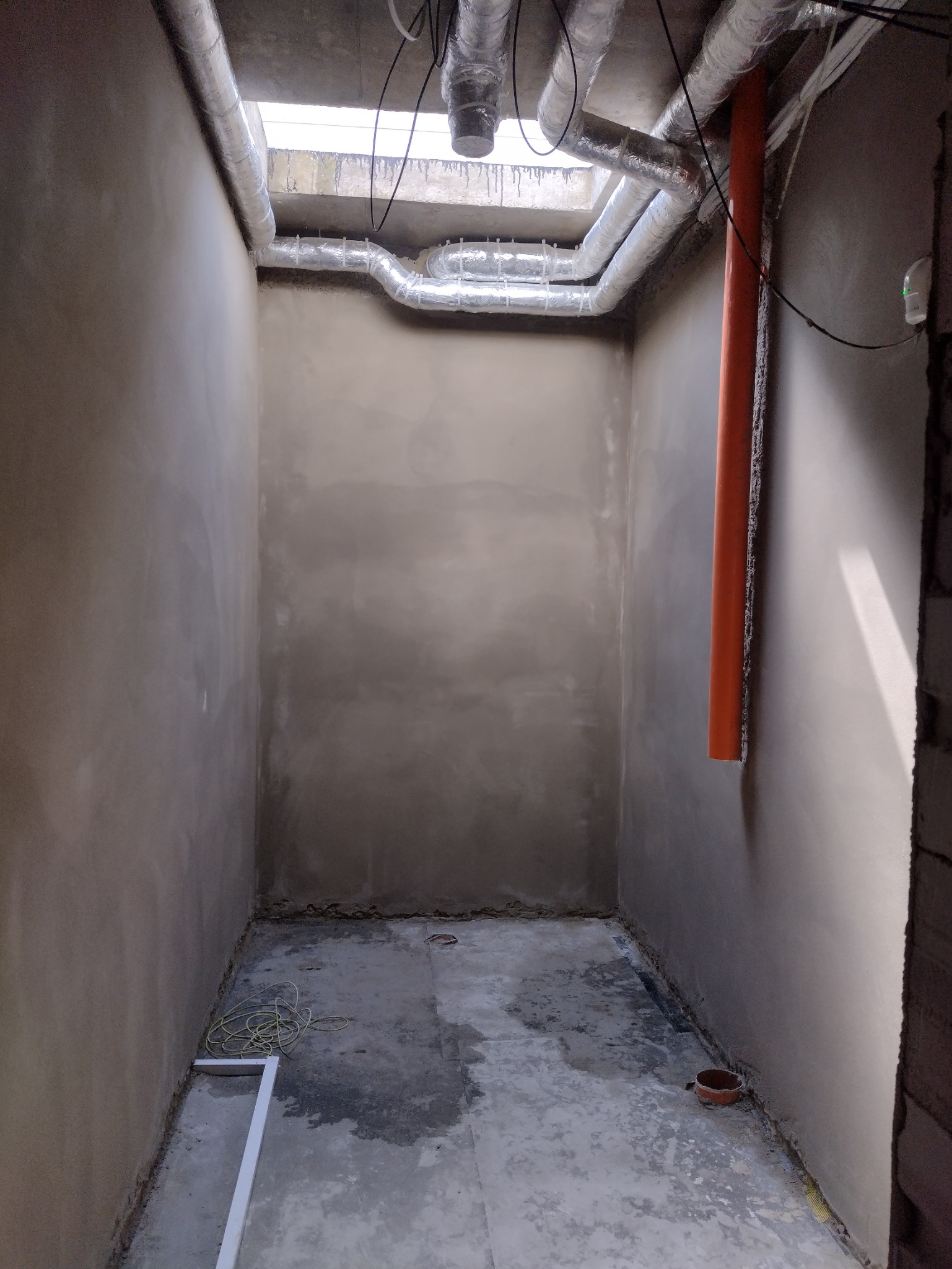 Wnętrze pomieszczenia po tynkowaniu maszynowym, widoczne rury wentylacyjne pod sufitem, pomarańczowa rura na ścianie i szary, surowy beton na podłodze.