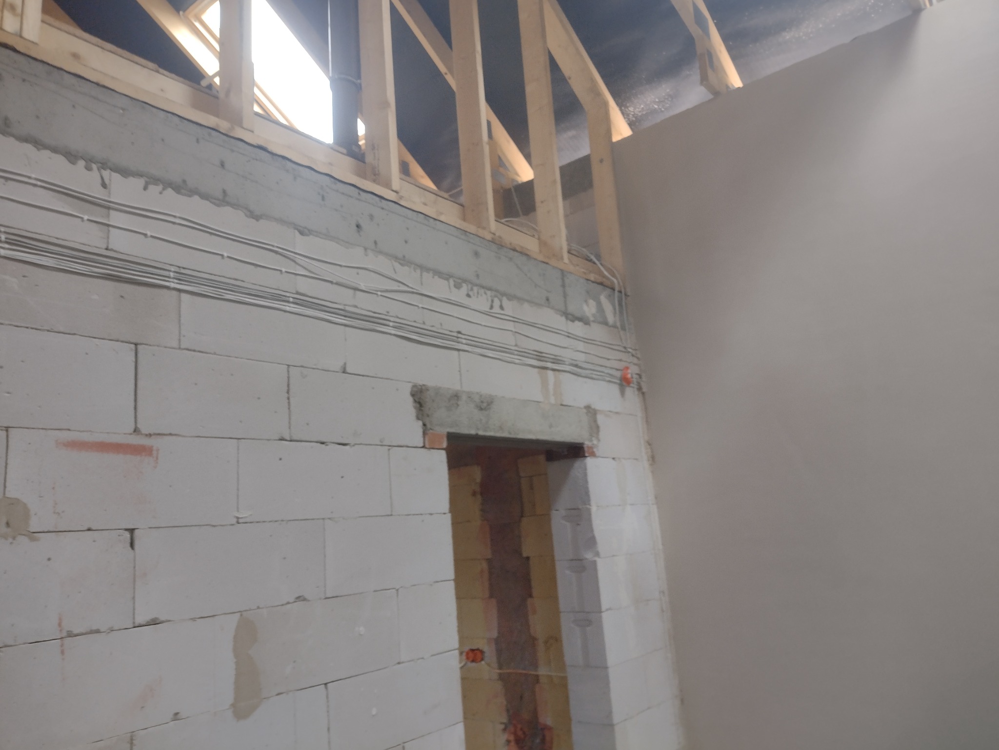 Surowa ściana z betonu komórkowego z widocznymi przewodami elektrycznymi, nadproże drzwiowe częściowo wykończone, fragment ściany wygładzonej, widoczna konstrukcja dachu z drewnianych belek.