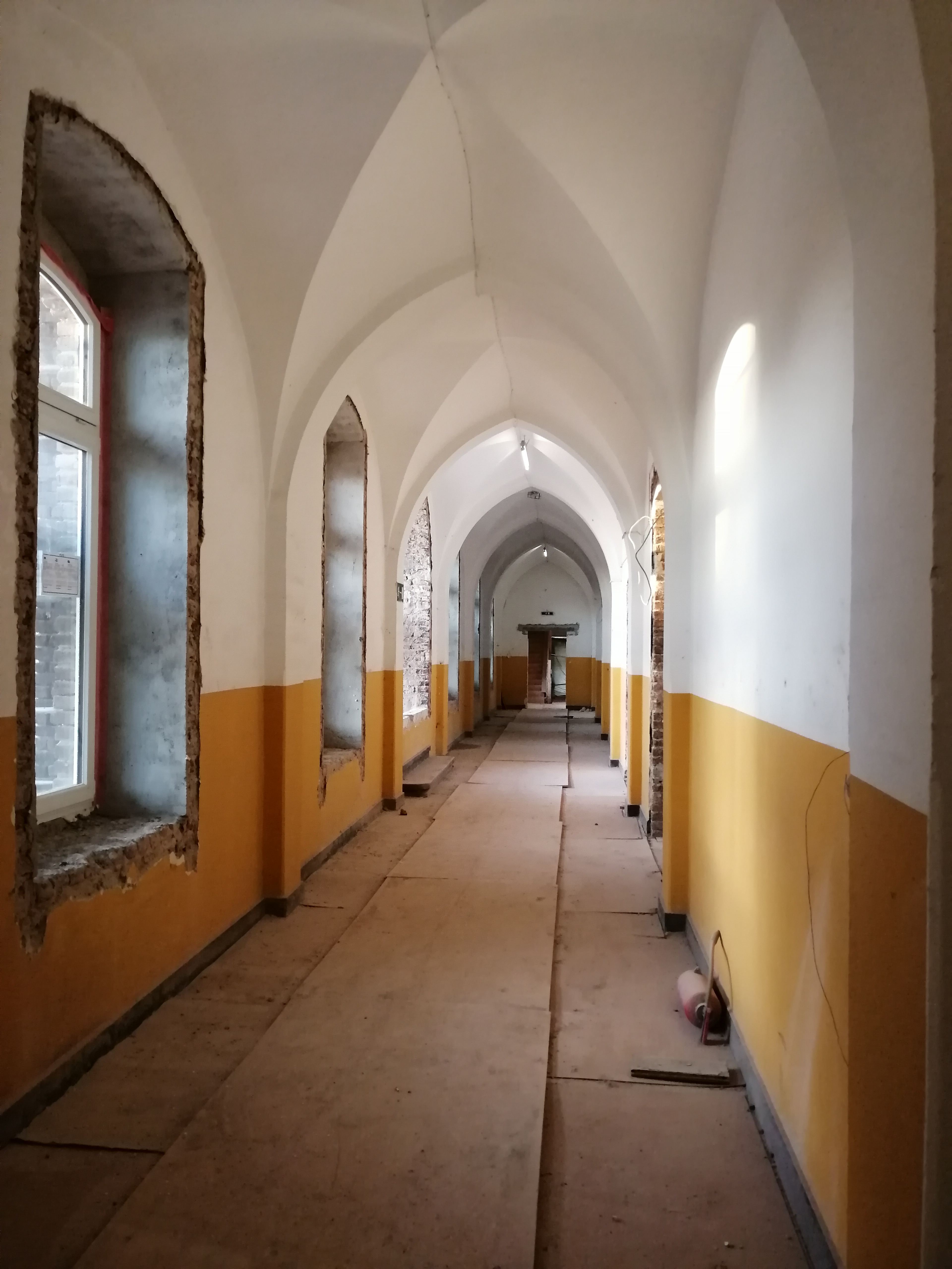 Długi korytarz z łukowym sklepieniem w trakcie remontu, widoczne przygotowane podłoże pod posadzkę, ściany częściowo pomalowane na żółto i biało, okna z surowymi obramowaniami.