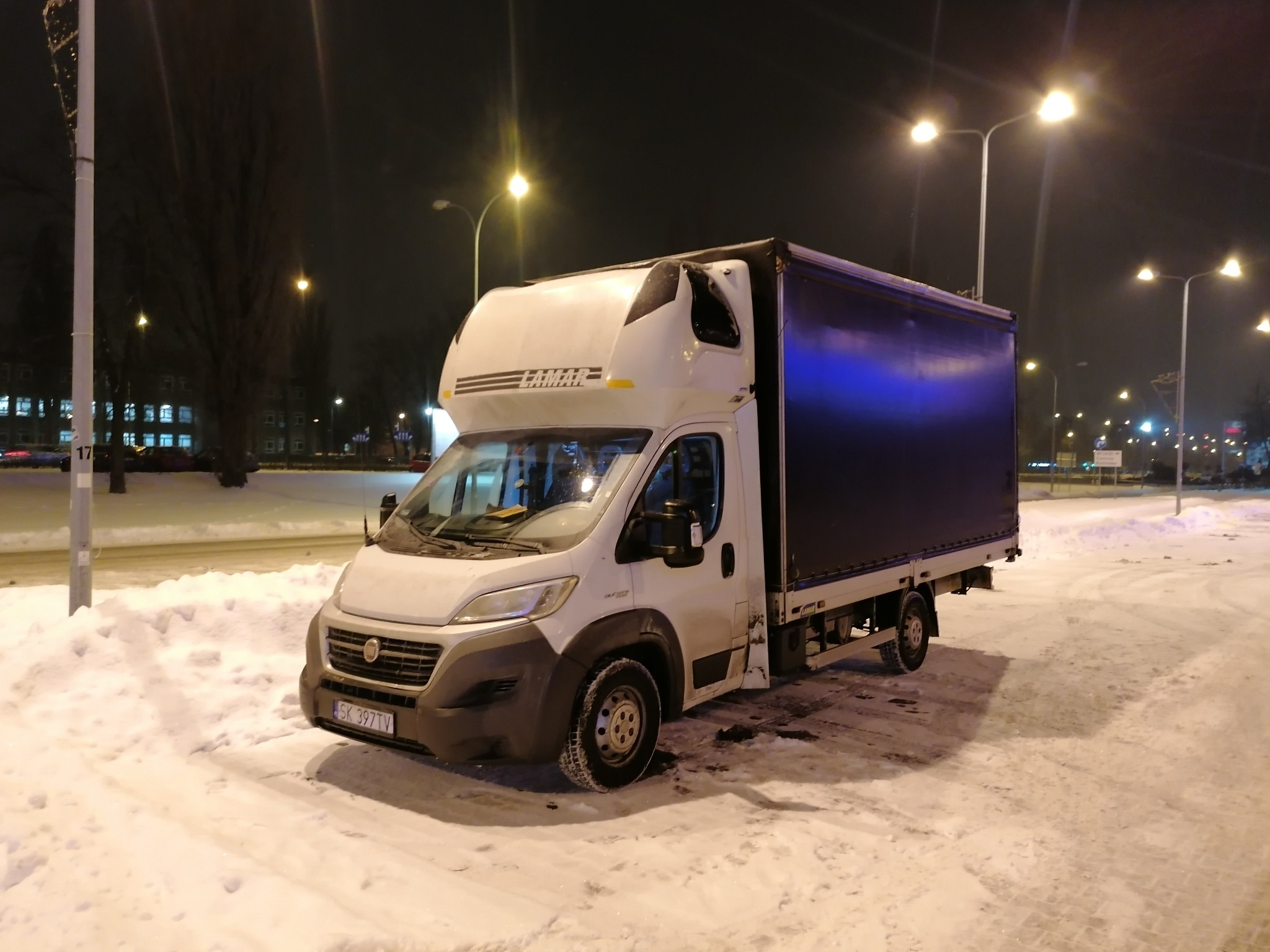 Biały samochód dostawczy z czarną plandeką zaparkowany na ośnieżonym parkingu nocą, oświetlony latarniami ulicznymi.