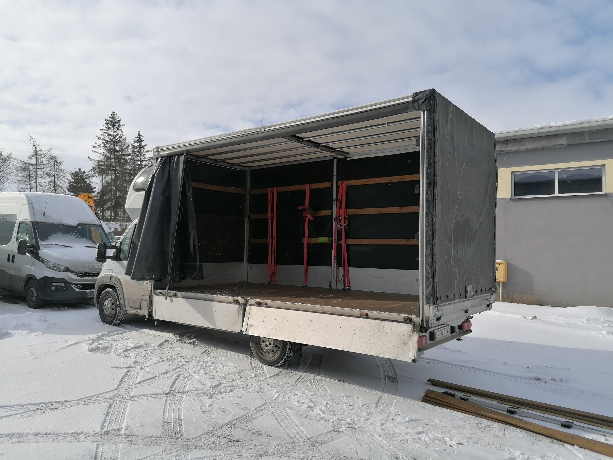 Otwarty samochód dostawczy z plandeką, widoczne pasy mocujące wewnątrz, zaparkowany na śniegu, obok budynek i inny dostawczak.
