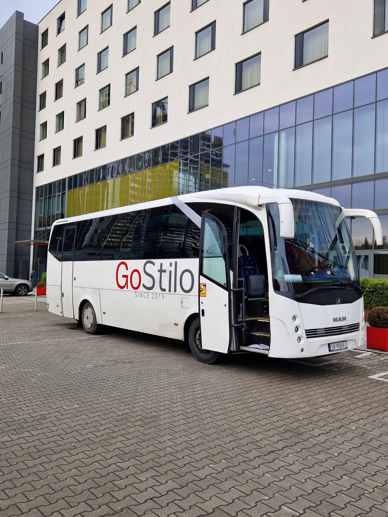 Biały autokar MAN z otwartymi drzwiami, oznaczony logo 'GoStilo SINCE 2019', zaparkowany przed nowoczesnym budynkiem z dużą ilością okien.