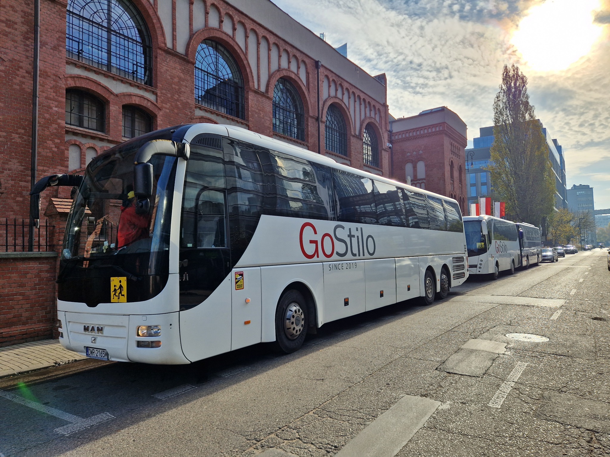 Biały autokar GoStilo zaparkowany na ulicy z kostki, w tle budynek z czerwonej cegły z dużymi oknami w kształcie łuku i rząd innych autokarów tej samej firmy.