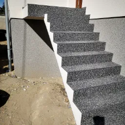 Zewnętrzne schody wejściowe wykończone szarą żywicą kamienną, z widoczną strukturą kruszywa, prowadzące do drzwi wejściowych budynku o jasnej elewacji.