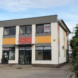 Fasada dwukondygnacyjnego budynku biurowego z oknami wystawowymi prezentującymi produkty i logo firmy, z elementami dekoracyjnymi w kolorach szarym, żółtym i czerwonym.