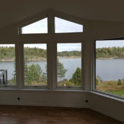 Jasne wnętrze z dużymi oknami z widokiem na jezioro i las. Widoczne drewniane podłogi i białe ściany z listwami. Charakterystyczne trójkątne okno na górze. Na zewnątrz widoczny budynek z czerwonymi...