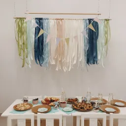 Stół z przekąskami i dekoracjami z wstążek w pastelowych kolorach, przygotowany na przyjęcie dla dzieci, z ciasteczkami, owocami i napojami w szklanych butelkach.