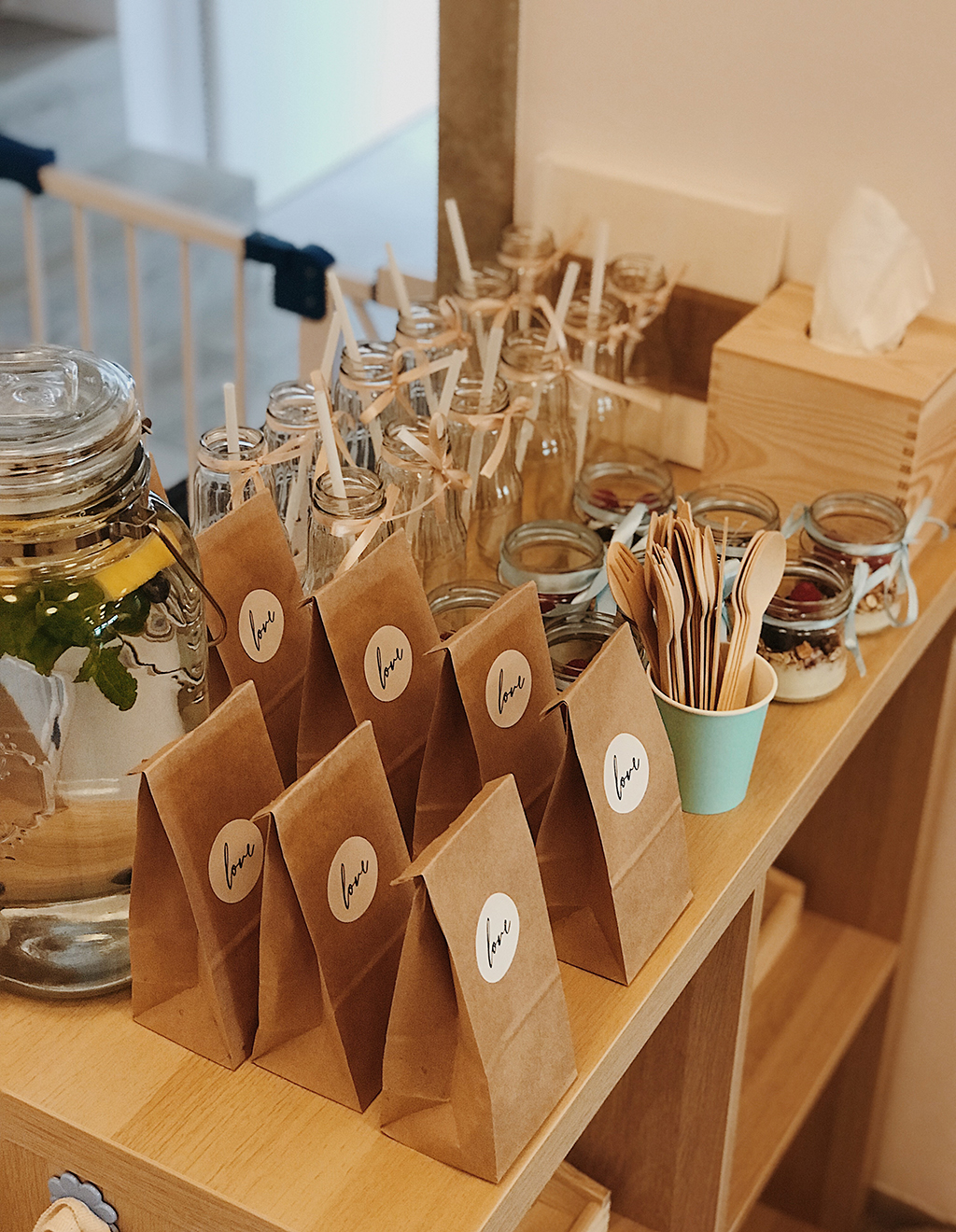 Przygotowany stół z przekąskami dla dzieci: szklane butelki ze słomkami, słoiki z owocami i granolą, papierowe torebki z naklejką 'love' oraz drewniane sztućce w turkusowym kubku.