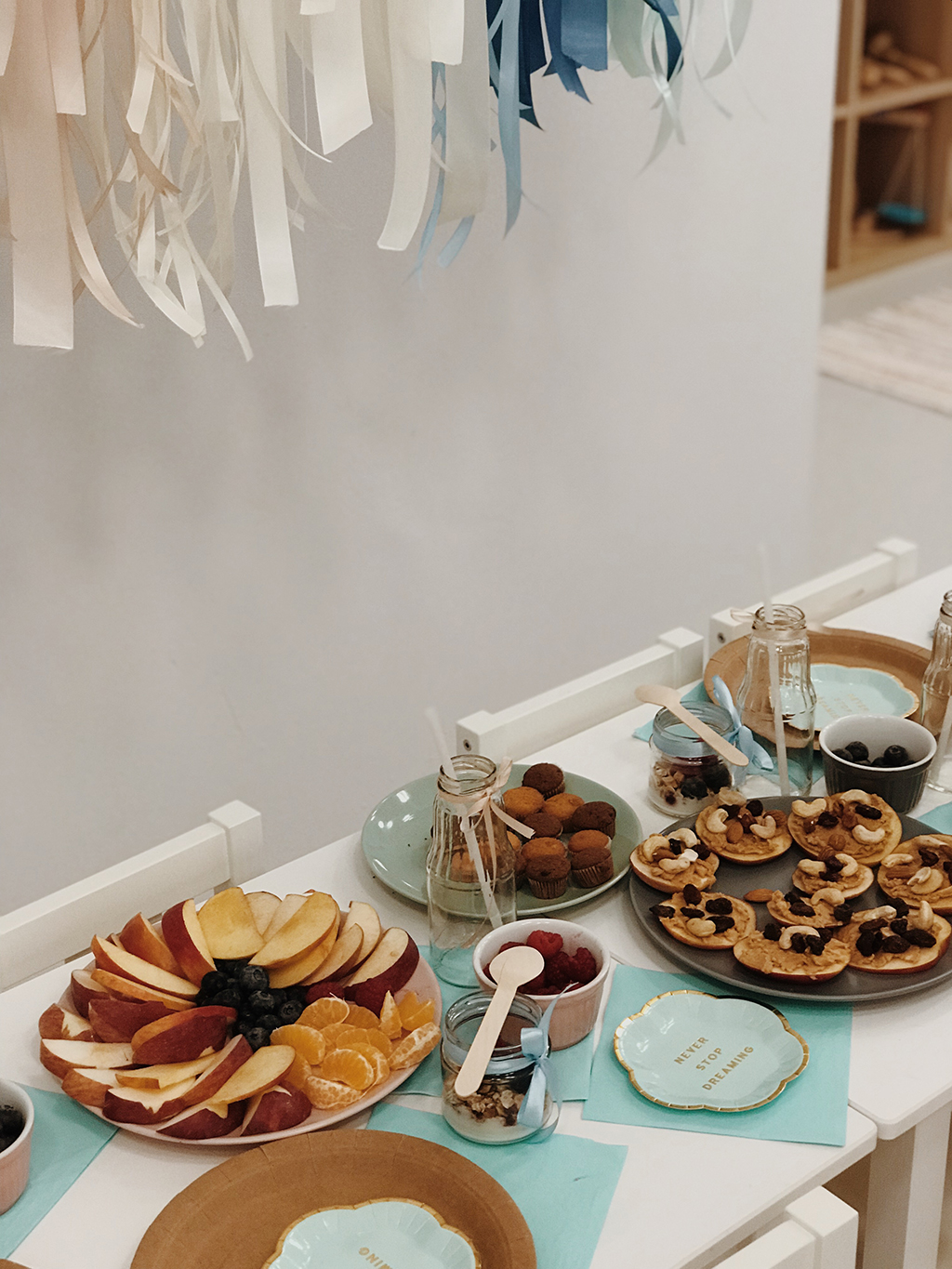 Stół z przekąskami dla dzieci: talerz z jabłkami, mandarynkami i jagodami, muffiny, mini-pizze z masłem orzechowym i bakaliami, musli w słoiczkach, w tle dekoracje z białych i niebieskich wstążek.