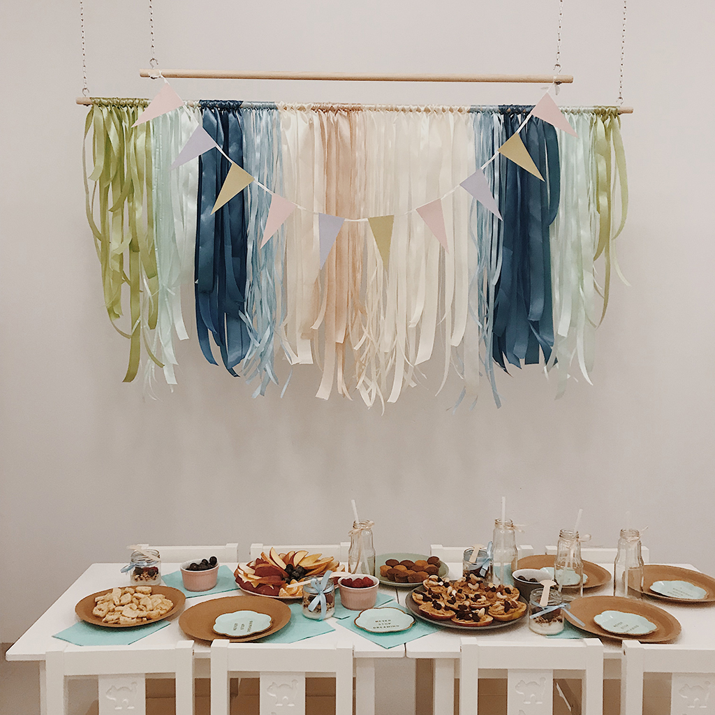 Stół z przekąskami i dekoracjami z wstążek w pastelowych kolorach, przygotowany na przyjęcie dla dzieci, z ciasteczkami, owocami i napojami w szklanych butelkach.