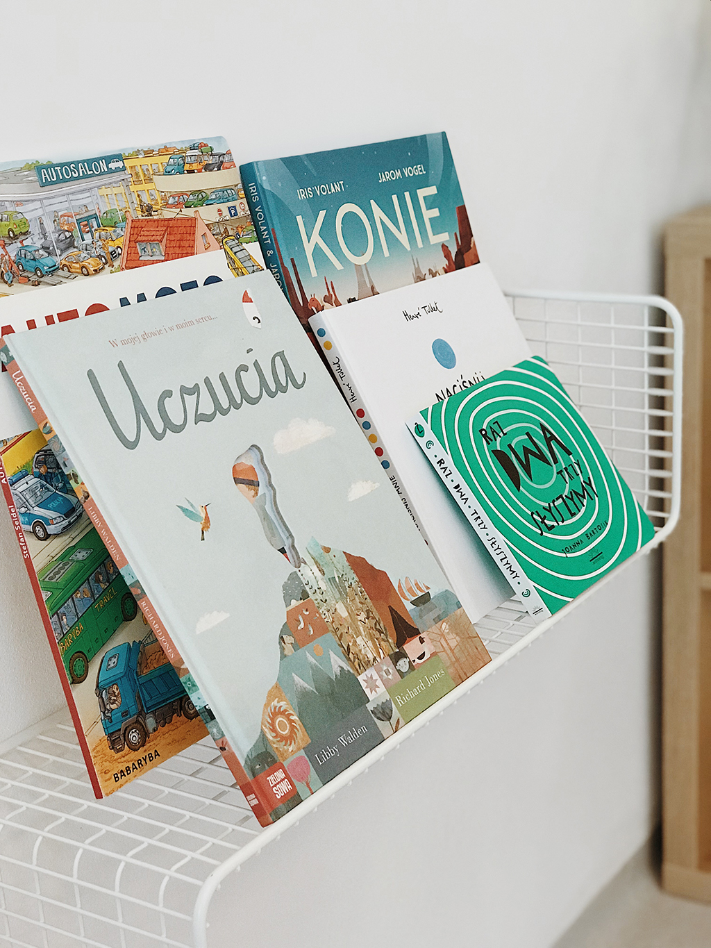 Książki dla dzieci ustawione w białym metalowym koszyku: 'Uczucia', 'Konie', 'Raz Dwa Trzy Słyszymy', 'Autosalon', z różnymi ilustracjami i okładkami.