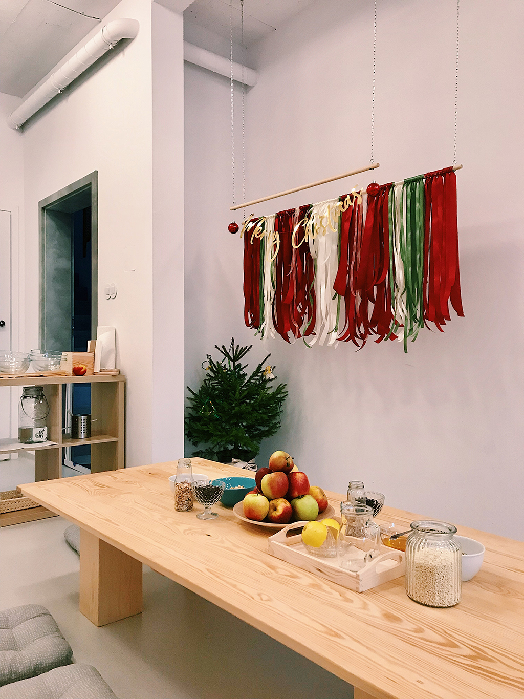 Sala żłobkowa w świątecznym klimacie: drewniany stół z owocami i słoikami, wisząca dekoracja z wstążek Merry Christmas, mała choinka w tle.