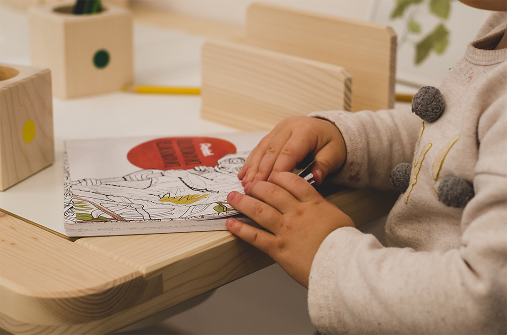 Dziecko koloruje kolorowankę na drewnianym stoliku, obok drewniane pojemniki na kredki z kolorowymi kropkami.