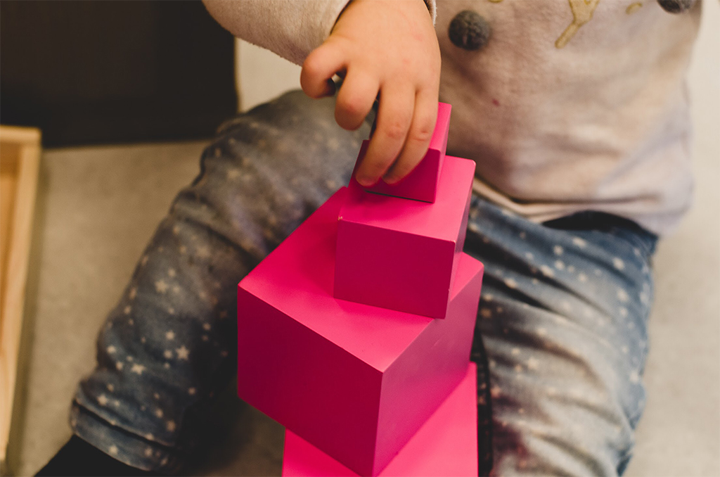 Dziecko układa piramidę z różowych drewnianych klocków o różnych rozmiarach, dżinsy w gwiazdki.