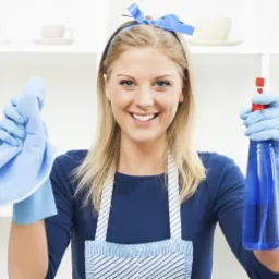 Uśmiechnięta blondynka w niebieskich rękawicach i fartuchu, trzymająca niebieską ściereczkę i spryskiwacz na tle białych półek.