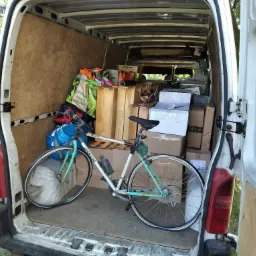 Wnętrze białego busa załadowane kartonami, drewnianymi elementami i rowerem Legnano z bidonem, widok z tyłu przez otwarte drzwi.