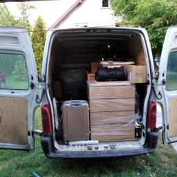 MoveTransport - Wnętrze białego busa z otwartymi drzwiami załadowane kartonami owiniętymi folią, walizką i torbami, widok z tyłu na trawnik i dom.