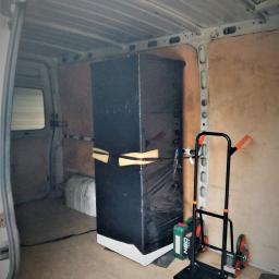 MoveTransport - Wnętrze furgonetki z zabezpieczonym meblem owiniętym folią, obok wózek transportowy z trzema kołami po każdej stronie i kanister z olejem. Ściany furgonetki wyłożone płytami ze sklejki.