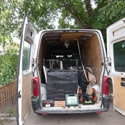 MoveTransport - Otwarty biały bus załadowany meblami owiniętymi w czarną folię stretch, narzędzia w skrzynkach i akcesoria do transportu, widok z tyłu na tle zieleni.