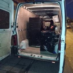 MoveTransport - Wnętrze białego busa załadowane meblami i kartonami zabezpieczonymi czarną folią stretch, widoczne otwarte tylne drzwi i fragment ulicy w tle.