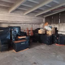 MoveTransport - Spakowane meble i kartony w czarnej folii stretch, przygotowane do transportu, składowane w magazynie o betonowych ścianach i suficie.