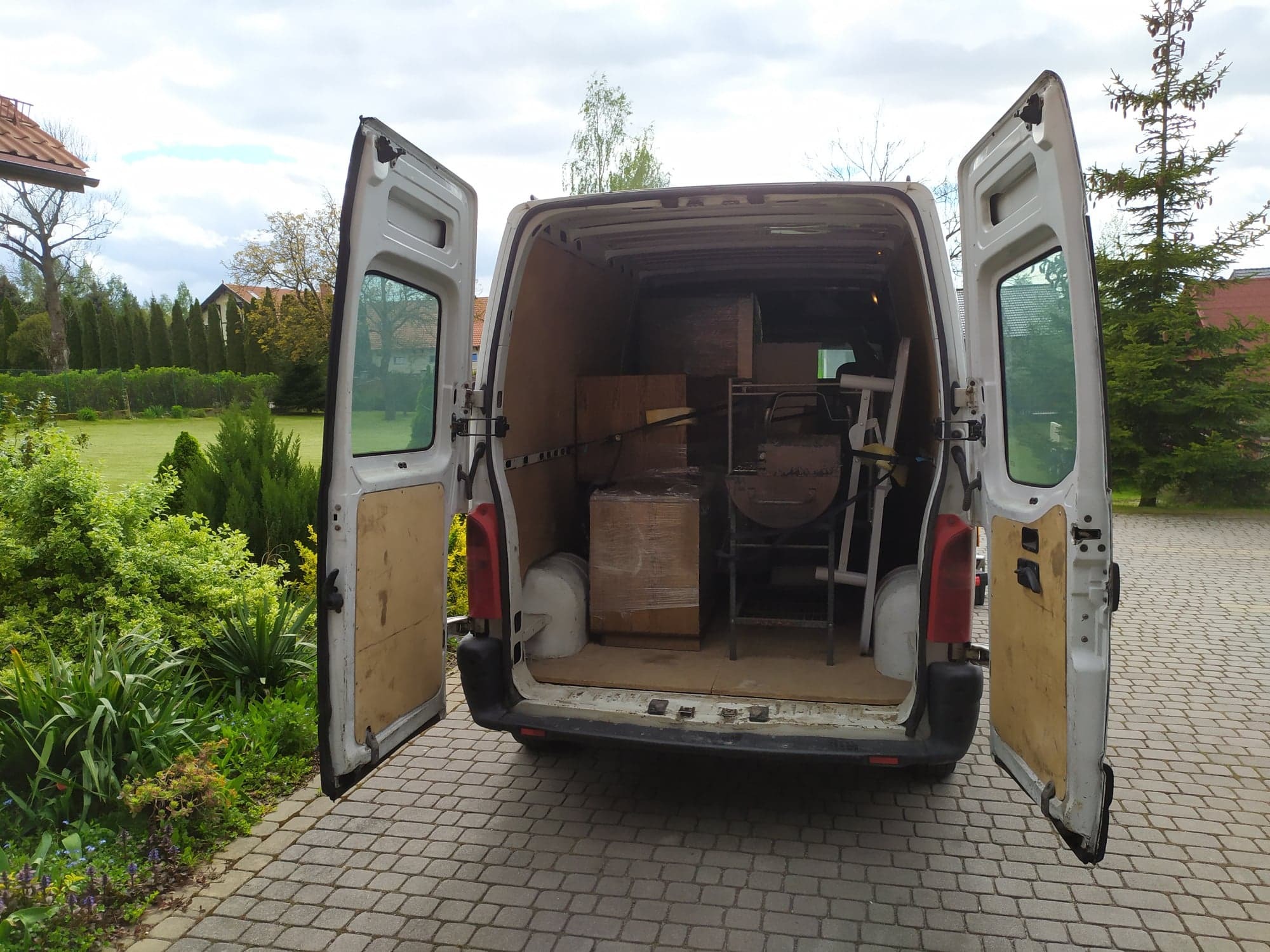 Wnętrze białego busa z otwartymi drzwiami załadowane kartonami owiniętymi folią stretch, krzesłami i metalowym grillem, widok z zewnątrz na brukowany podjazd i zieleń.