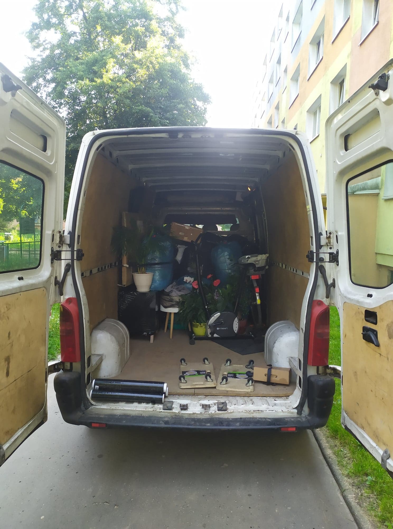 Otwarty tył białego vana załadowanego roślinami doniczkowymi, workami i sprzętem do ćwiczeń, widok z zewnątrz na tle bloku mieszkalnego i drzew