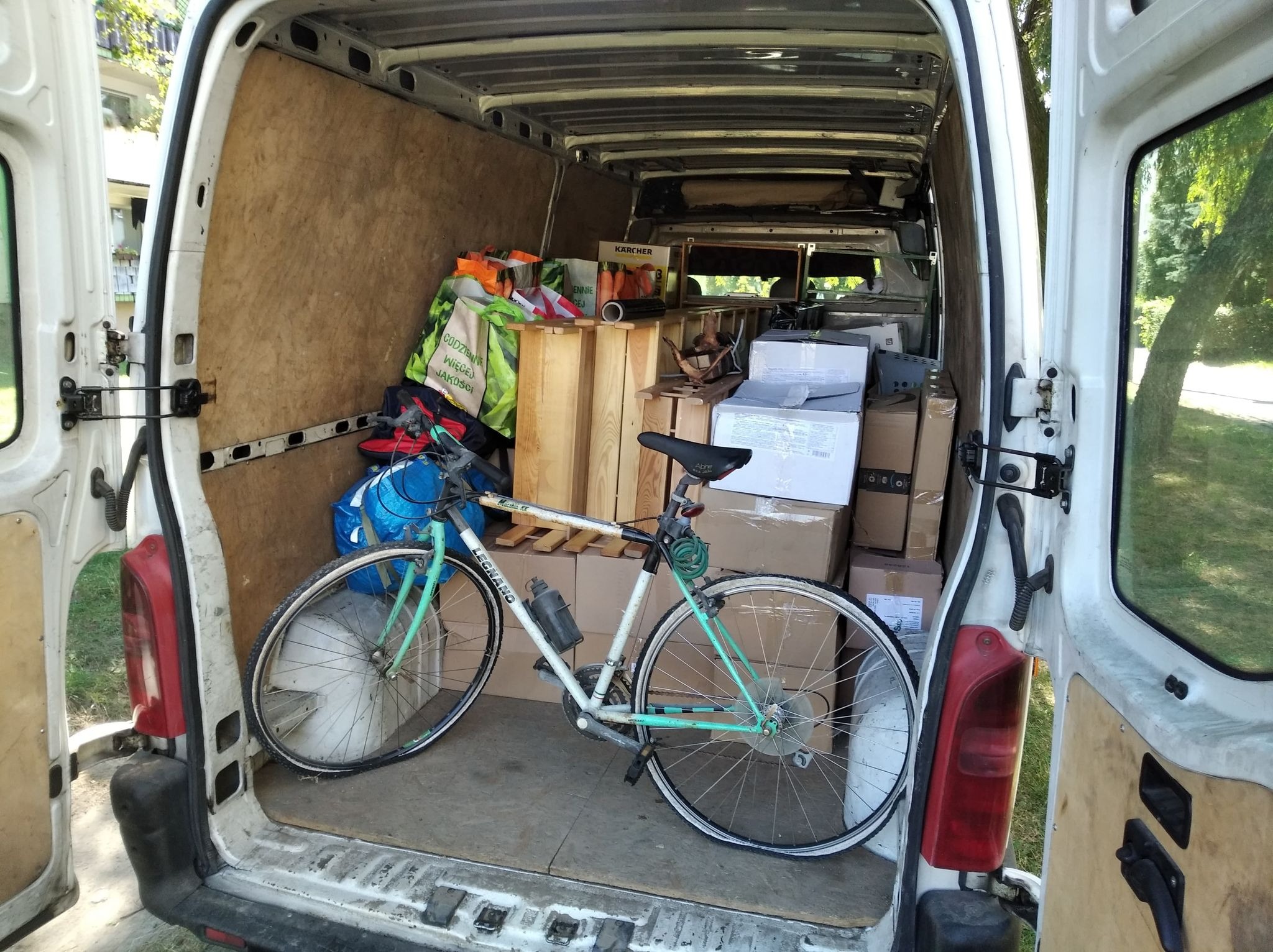 Wnętrze białego busa załadowane kartonami, drewnianymi elementami i rowerem Legnano z bidonem, widok z tyłu przez otwarte drzwi.